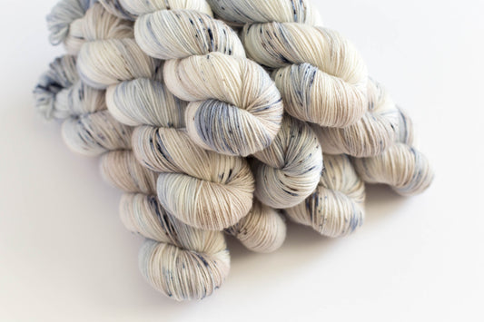 Farmhouse knitting yarn. Angled pile of variegated fingering yarn skeins in a soft off-white, cream, and slate blue-gray colorway, photographed on a clean white background.