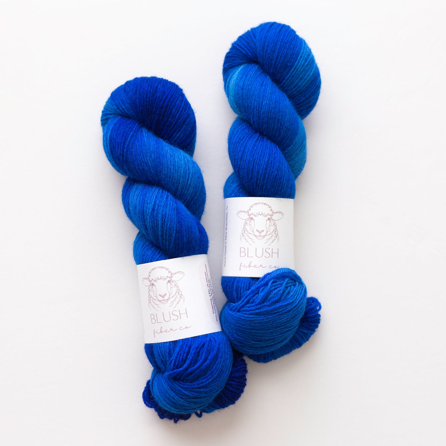 Two skeins of hand dyed variegated blue yarn with white paper labels reading 'BLUSH  fiber co.' on a clean white background.